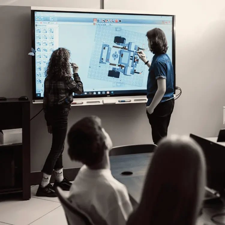 Students using an interactive panel to collaborate on a 3D design project in a classroom setting, with peers observing.