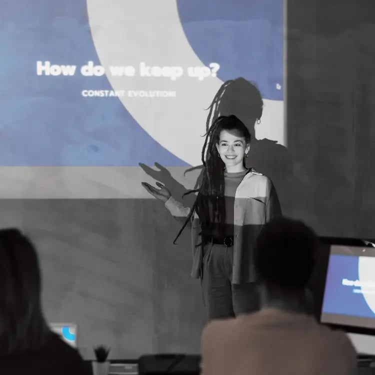 Teacher presenting in front of a projected screen during a classroom session, with students viewing the presentation on devices. Teacher presenting in front of a projected screen during a classroom session, with students viewing the presentation on devices.
