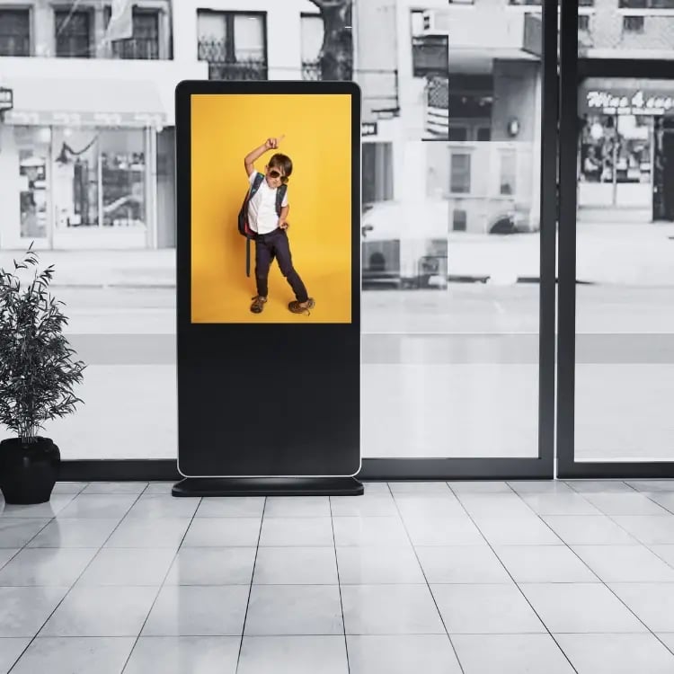 Freestanding digital signage display in a school entrance showing a student with backpack on a bright yellow background, used for welcoming messages and school communication.